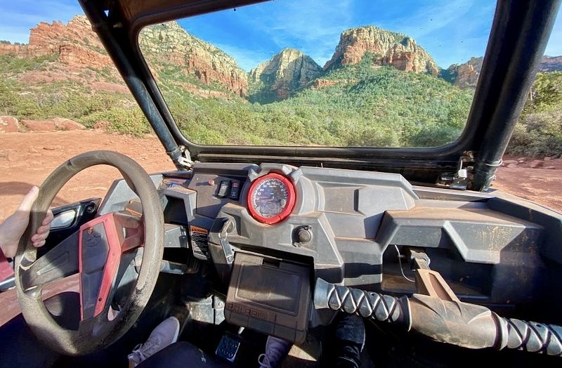 Breathtaking off-road trails weaving through the red rock formations of Sedona, vibrant desert landscape under a bright blue sky, rugged UTV vehicles navigating rocky paths, distant mountain ranges, cacti and wildflowers dotting the scenery, shadow play on the ground from sunlit cliffs.