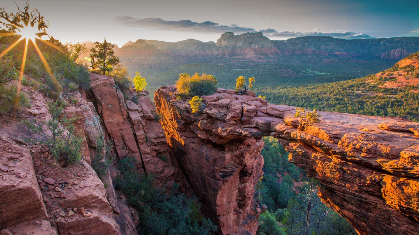 A picturesque landscape of Sedona showcasing vibrant red rock formations, a winding off-road trail perfect for UTVs, and a recreational vehicle parked at the side. The scene is bathed in warm sunlight, highlighting the contrasting green vegetation against the striking red rocks, with a clear blue sky overhead and distant mountains framing the horizon.